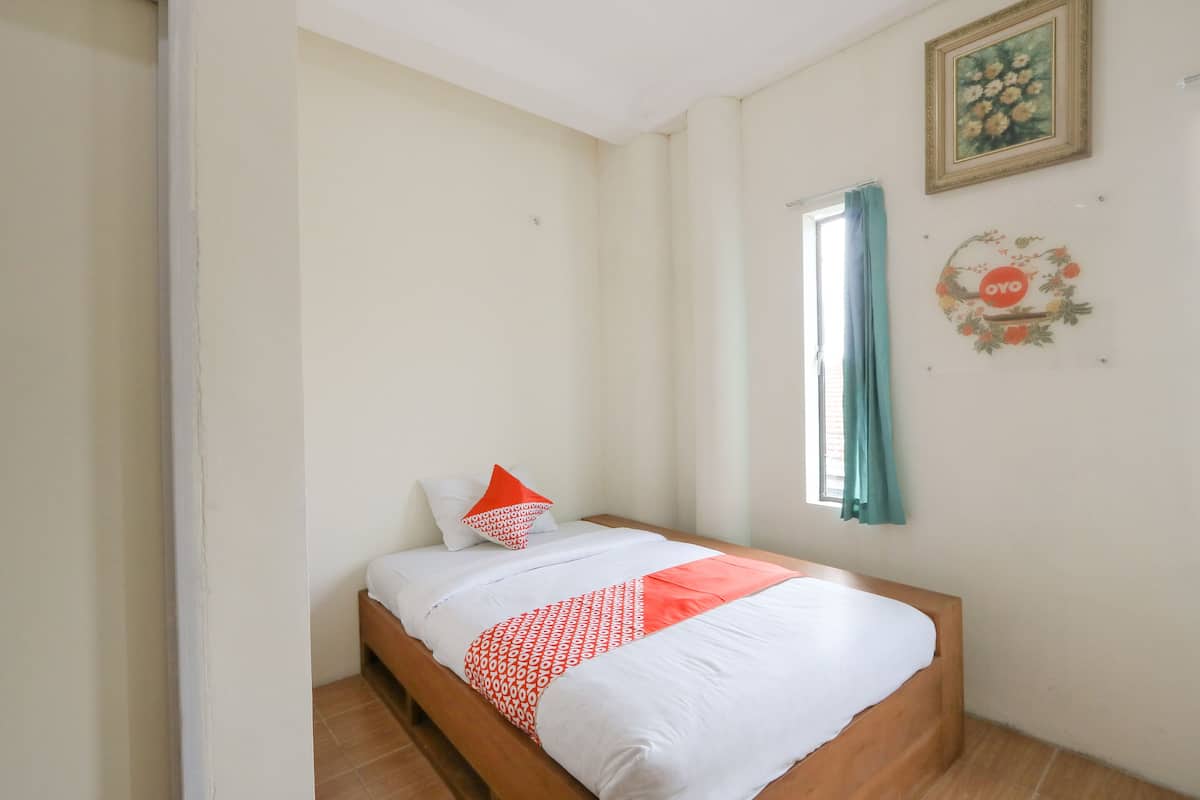 A bedroom with a wooden bed, white bedding, a red and white pillow, a window with a curtain, and a wall decoration.