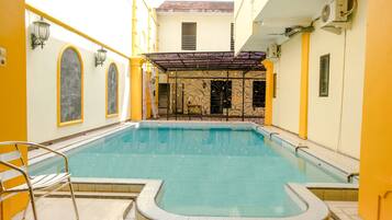 A swimming pool with a tiled deck, a covered seating area, and a yellow and white building facade.