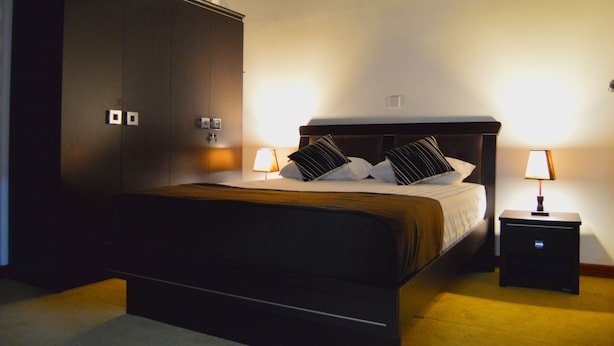 Hotellihuone, jossa on suuri vaatekaappi, puinen päätyosaan varustettu sänky, kaksi yöpöytävalaisinta ja matolla peitetty lattia.