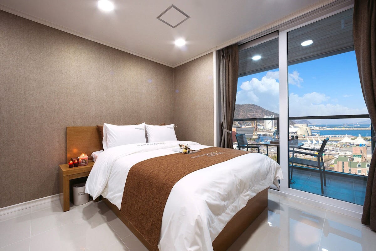 Quarto de hotel moderno com uma cama grande, uma mesa de cabeceira com velas e vista para a cidade através de amplas janelas.