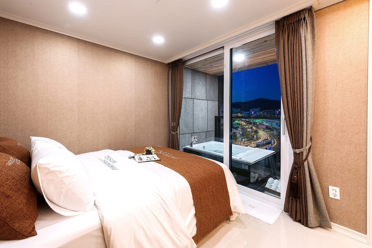 Quarto com cama, mesa de cabeceira, janela com vista para a cidade e porta deslizante.