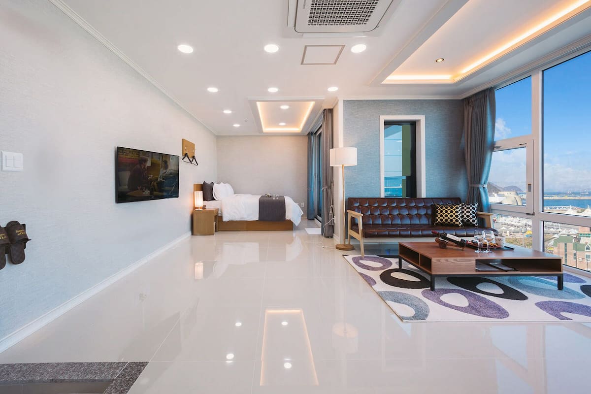 Quarto de hotel moderno com uma cama, um sofá, uma televisão e uma janela grande com vista para a cidade.