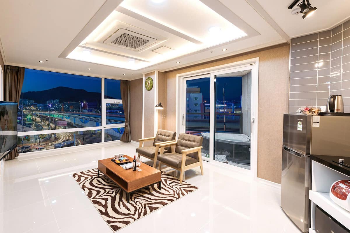 Uma sala de estar moderna com vista para a cidade, uma televisão de tela plana, uma mesa de centro e uma cozinha com geladeira.