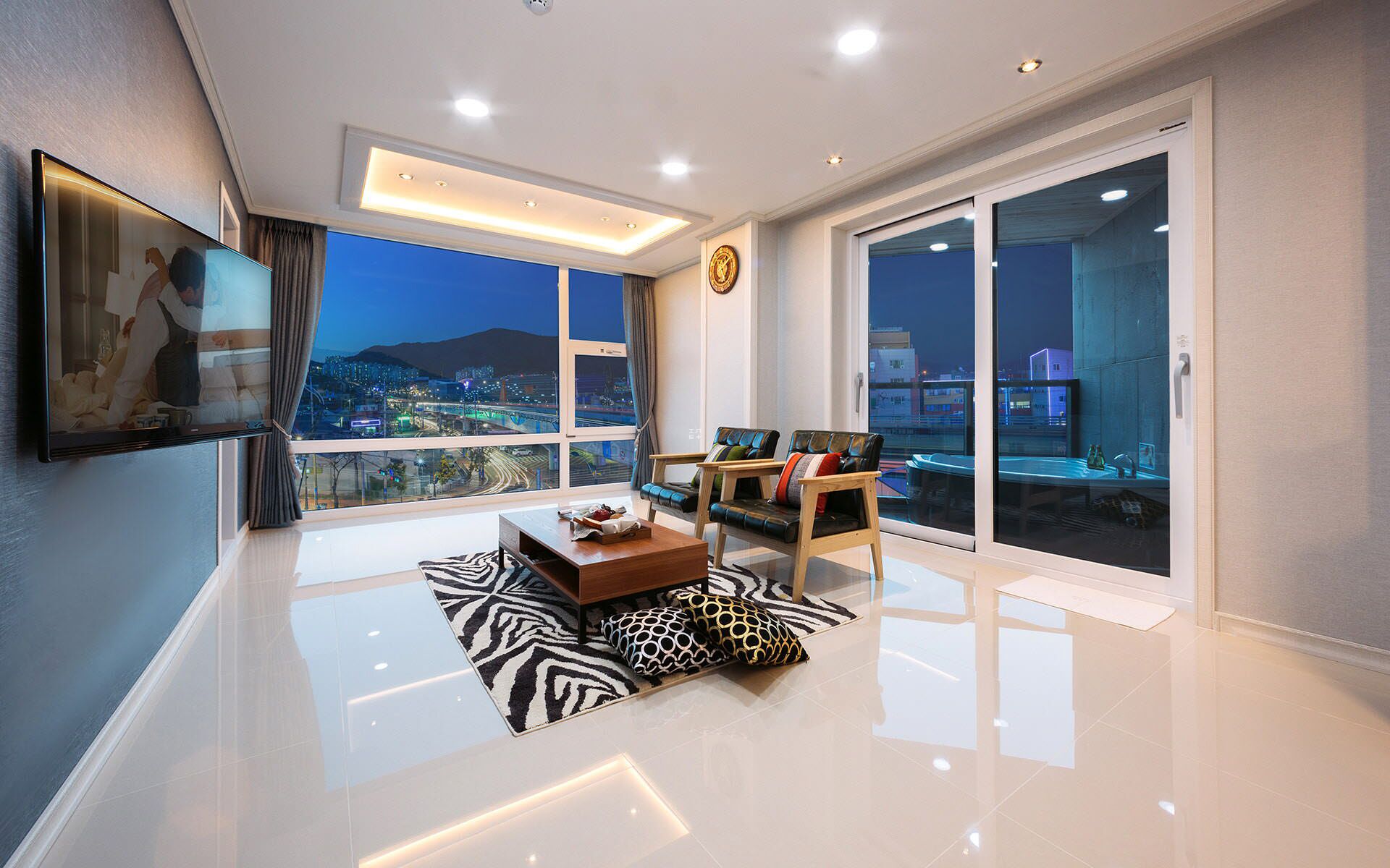 A modern living room with a large TV, a sofa, a coffee table, and a view of a cityscape at dusk.