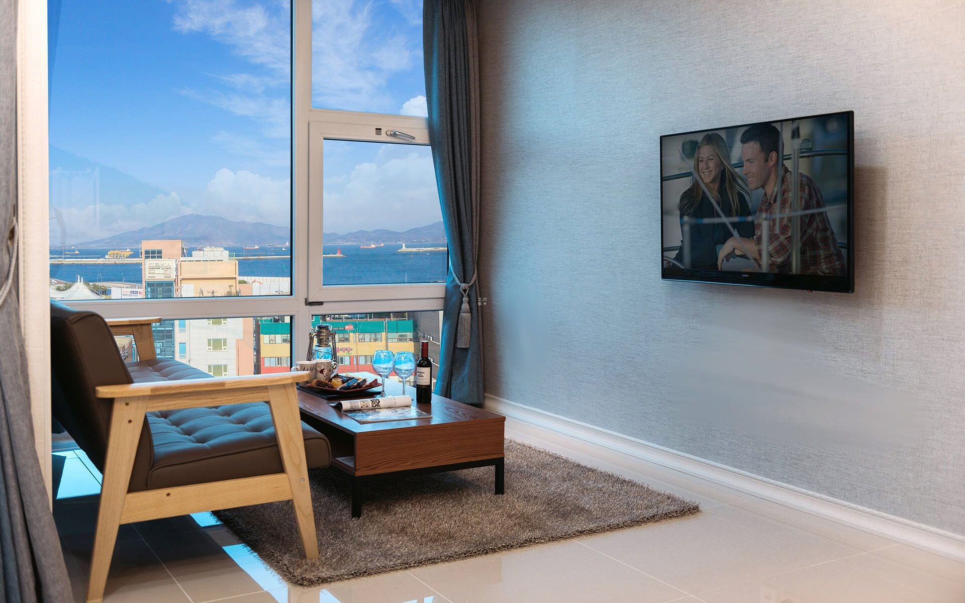 A modern living room with a large window, a sofa, a coffee table, and a TV mounted on the wall.