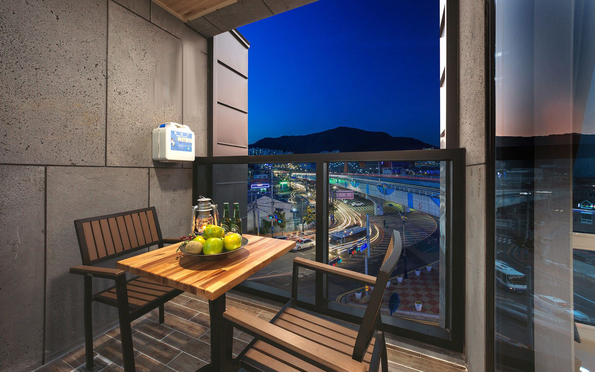 A balcony with a wooden table, chairs, a lantern, and a bowl of fruit. A cityscape with a bridge and lights is visible through the window.