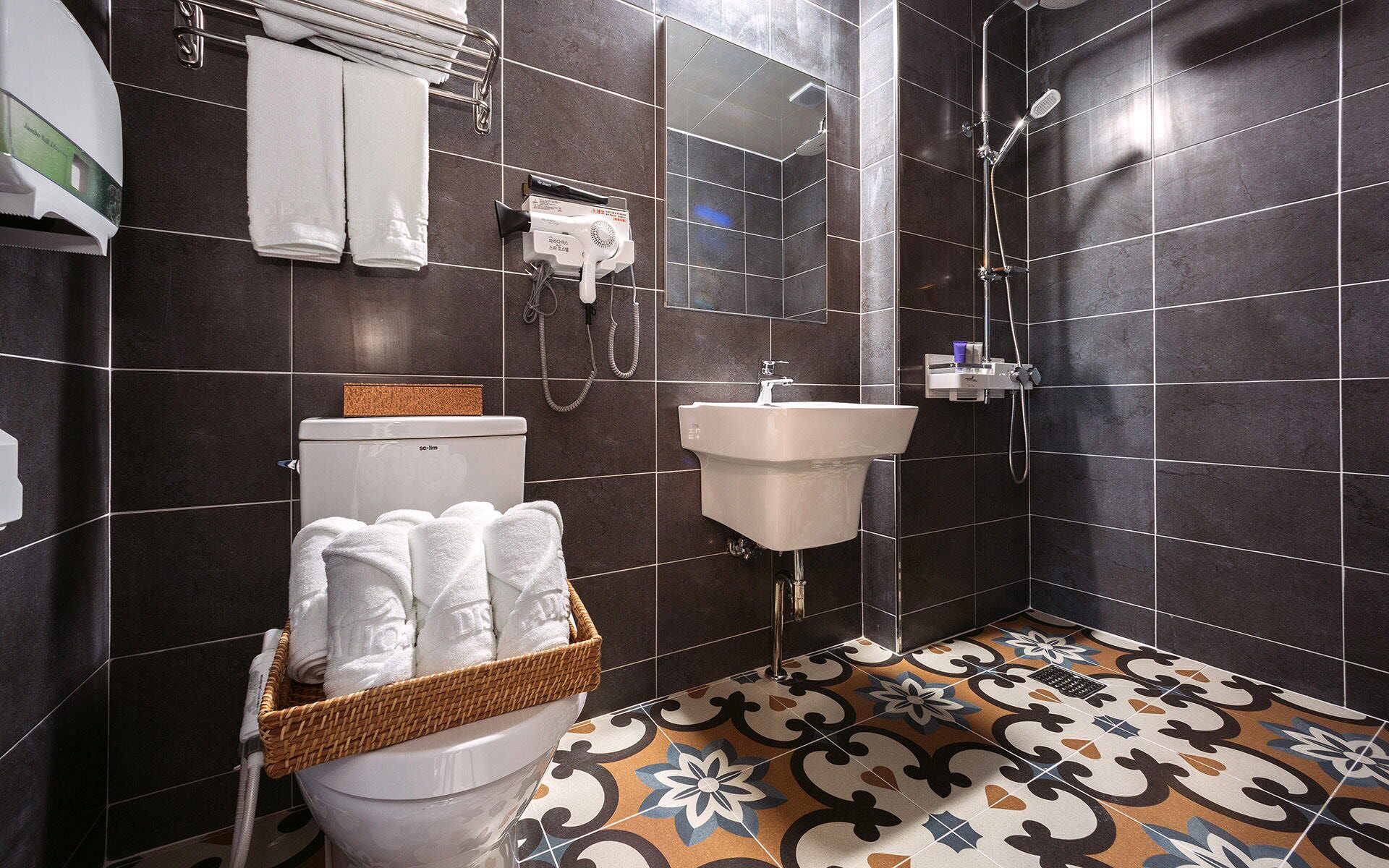 A bathroom with dark tiled walls, a white sink, a shower, and a basket of white towels.