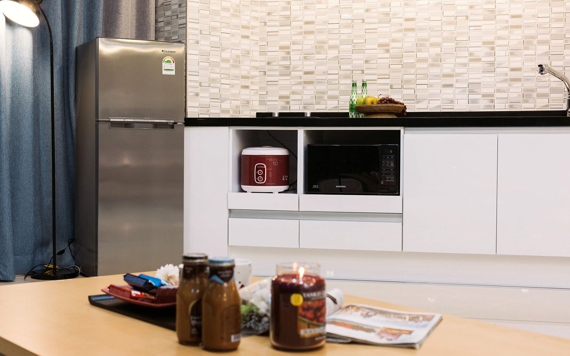A kitchen with a stainless steel refrigerator, a rice cooker, and a microwave. There are jars on the counter and a candle.
