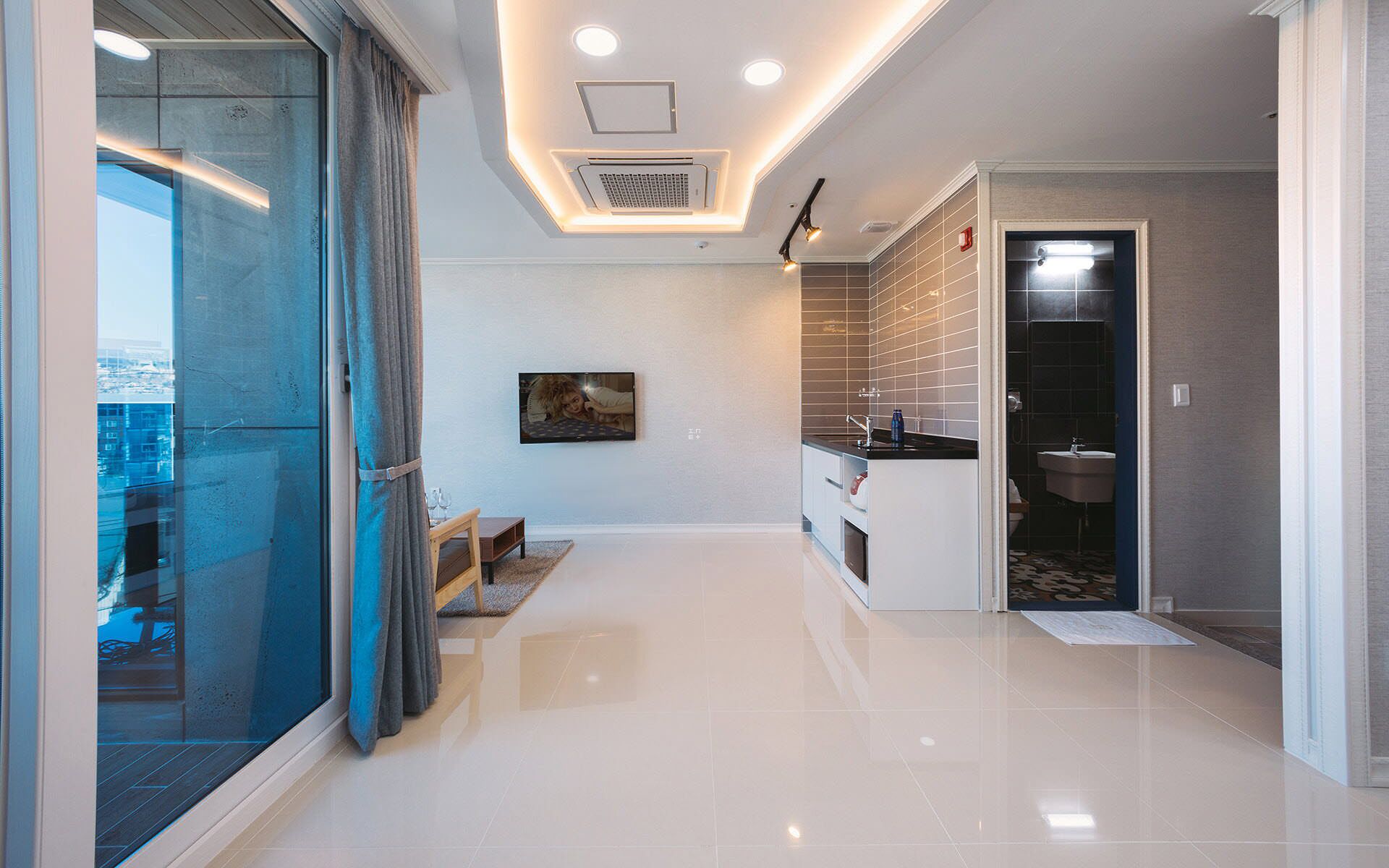 A modern hotel room with a glass door leading to a balcony, a small seating area, and a kitchenette.
