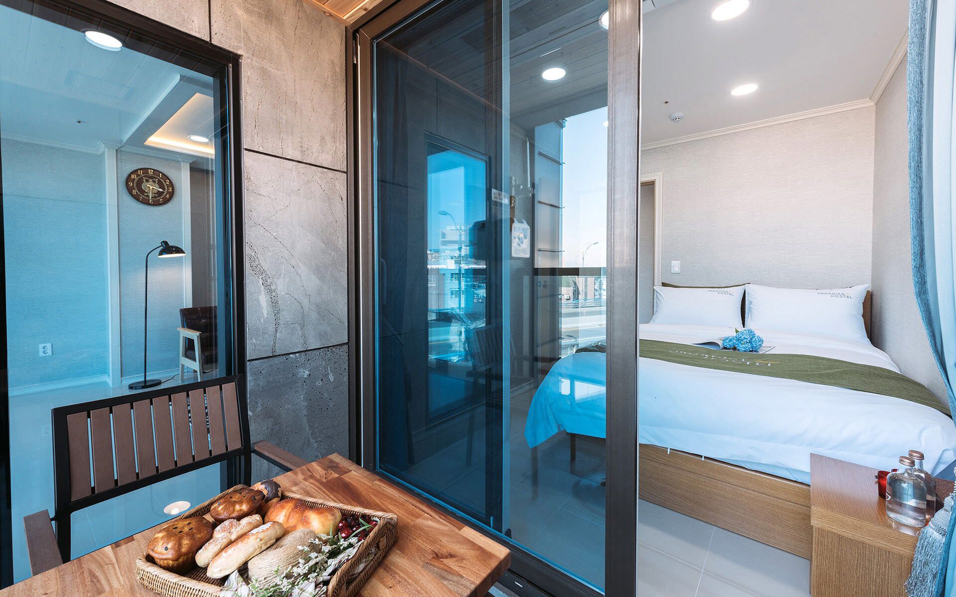 A modern hotel room with a bed, a wooden table with bread, and a view through a glass door.