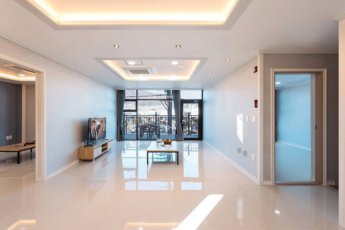Um hall de hotel espaçoso com design moderno, apresentando uma grande porta de vidro que dá para uma área externa, um espaço de estar com mesa e cadeiras, e uma televisão sobre um suporte de madeira.