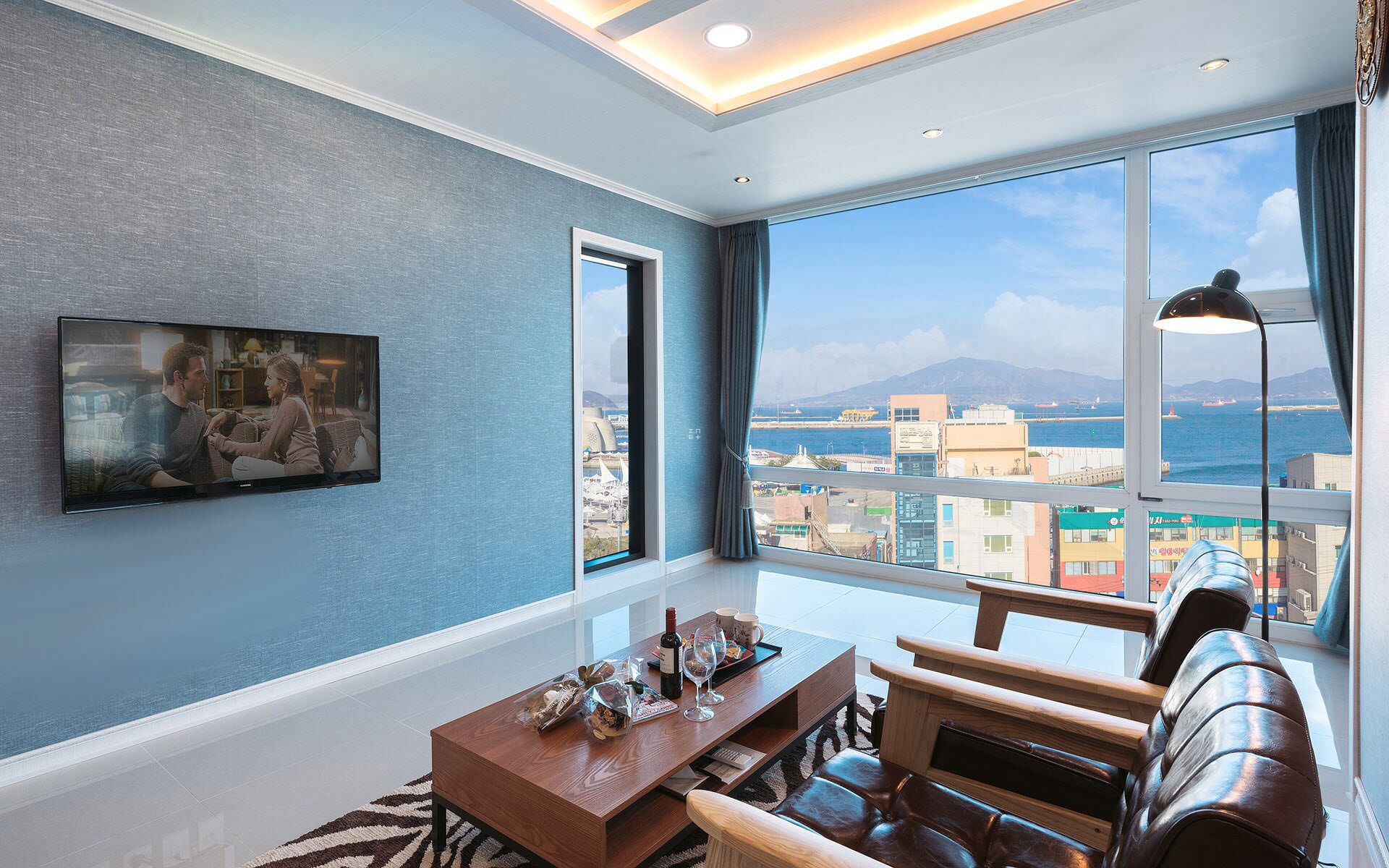 A modern living room with a large TV, a wooden coffee table, leather sofa, and a view of the city and sea.