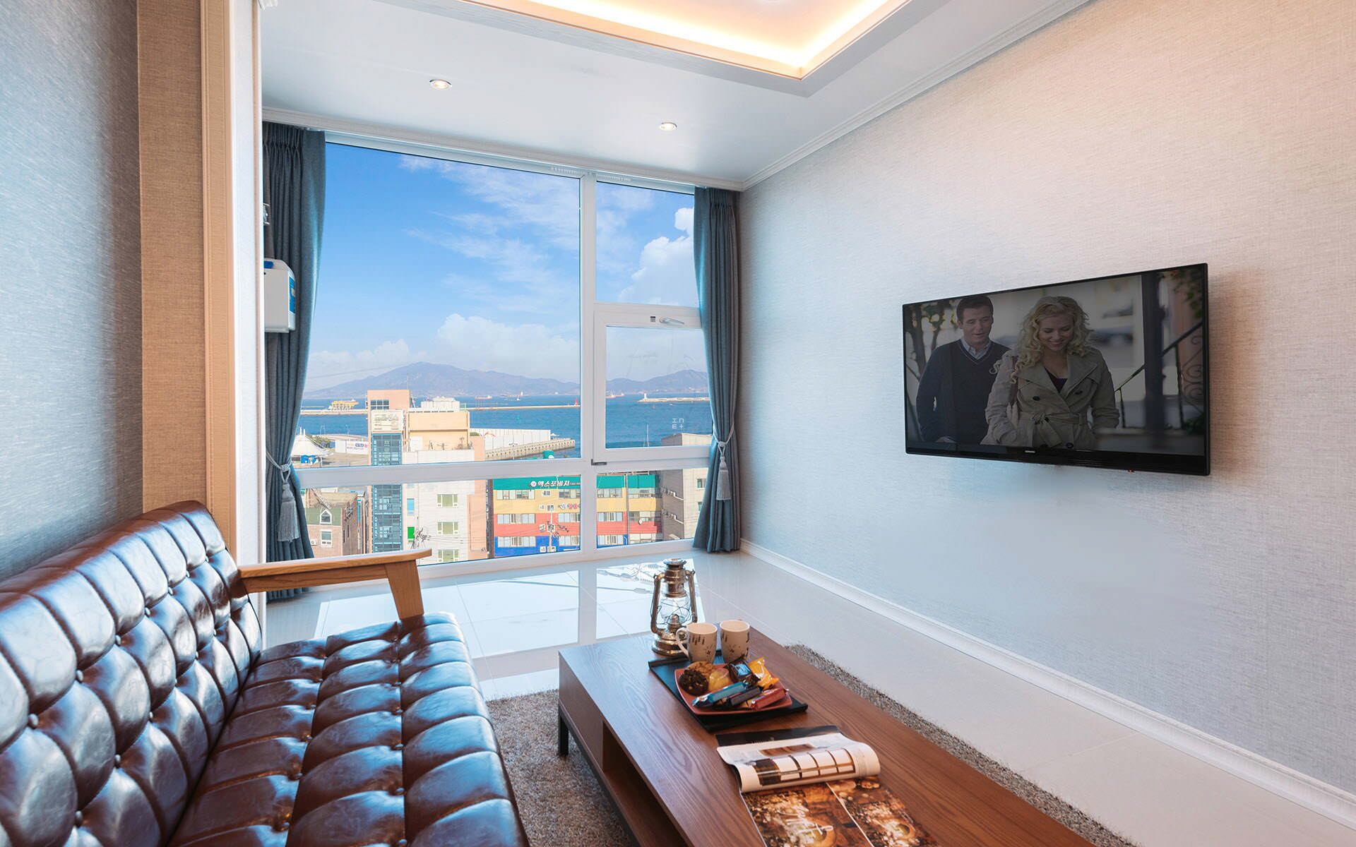 A modern living room with a leather sofa, a wooden coffee table, and a large window offering a city view.
