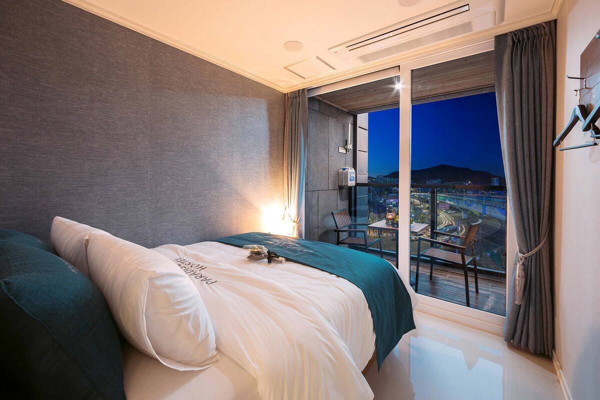 Quarto de hotel com cama, mesa de cabeceira, janela com vista para a paisagem urbana e varanda com cadeiras.