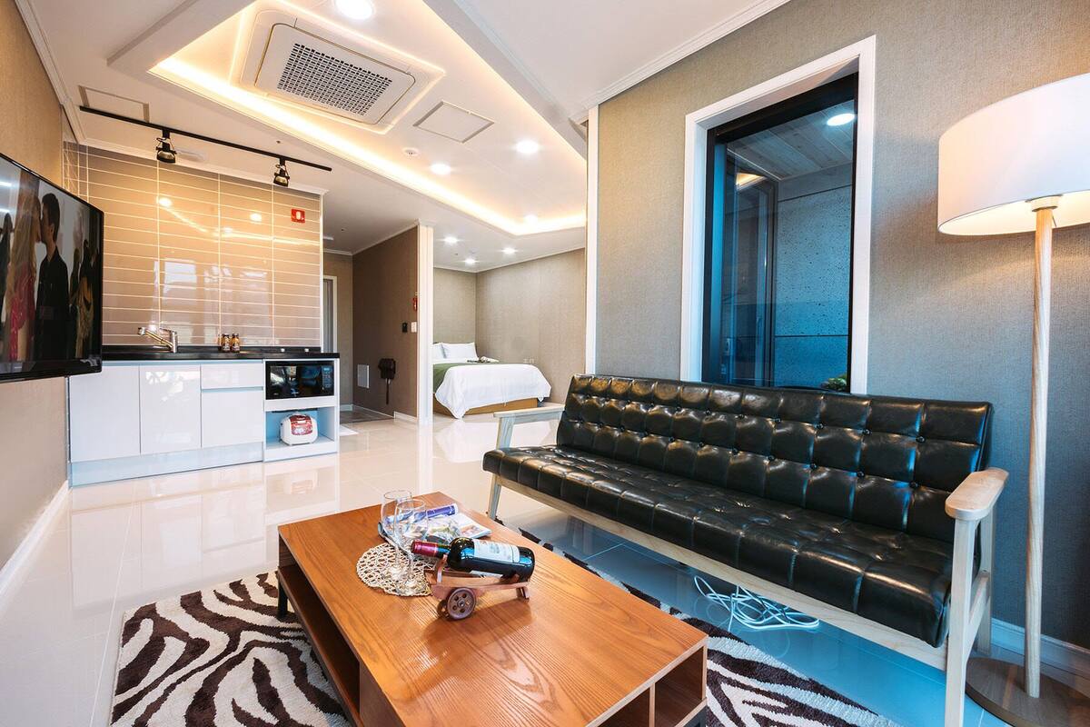 Quarto de hotel moderno com sofá, mesa de centro de madeira, televisão, área de cozinha e banheiro visível através de uma porta de vidro.