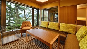 A modern living room with a wooden floor, a sofa with patterned cushions, a wooden coffee table, and large windows offering a view of trees and a body of water. - Biwako Mastunoura Bettei (Otsu)