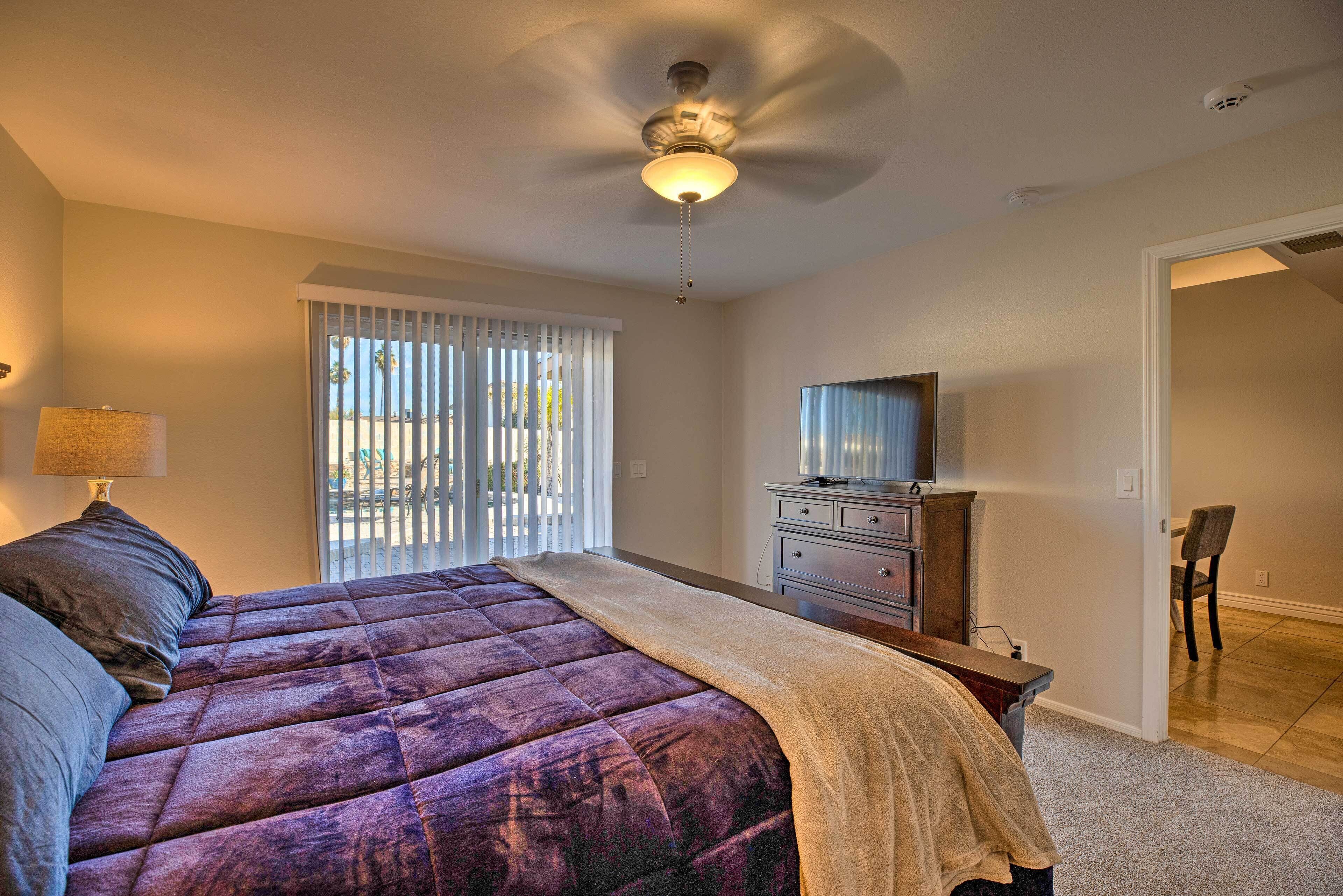 A bedroom with a large bed, a ceiling fan, a TV, and a window with blinds.