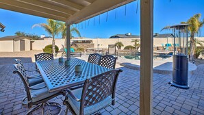 Un patio al aire libre con una mesa y sillas de metal, una pérgola arriba y vistas a una piscina y edificios al fondo.