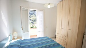A bedroom with a bed, a wardrobe, a nightstand with a lamp, and a view of a mountainous landscape through a window. - BOUGAINVILLEA PRIMO PIANO (Marciana)