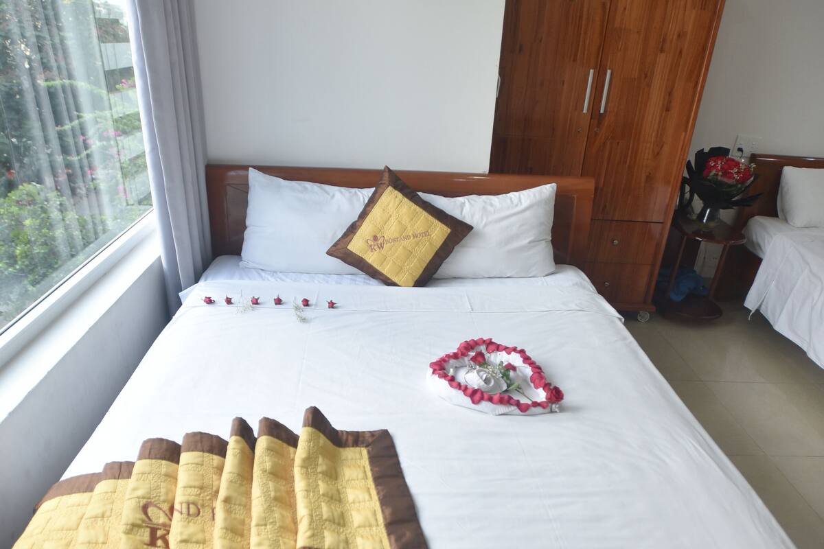 Una cama bien hecha con una colcha blanca y marrón, una almohada decorativa y un arreglo floral. Se ve un armario de madera al fondo.