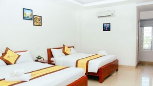 A hotel room with three beds, each with a wooden frame and white bedding. There are wall-mounted air conditioning units and framed pictures on the walls.