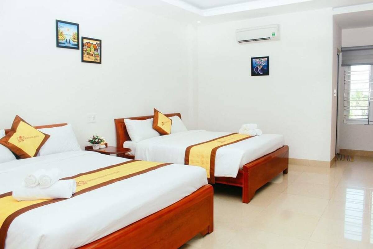 Habitación de hotel con tres camas, cada una con estructura de madera y ropa blanca. Hay unidades de aire acondicionado fijadas a la pared y cuadros enmarcados.