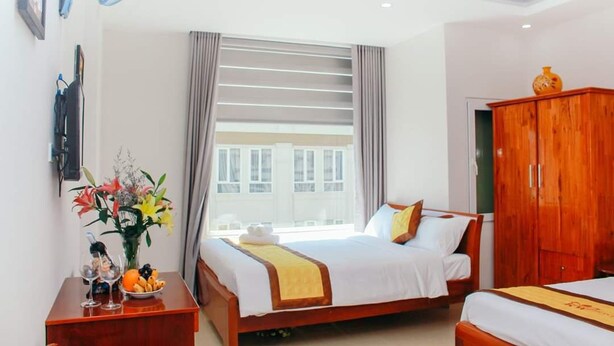 A hotel room with two beds, a wooden wardrobe, a desk with a vase of flowers, and a window with blinds.