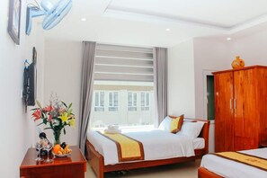A hotel room with two beds, a wooden wardrobe, a desk with a vase of flowers, and a window with blinds.