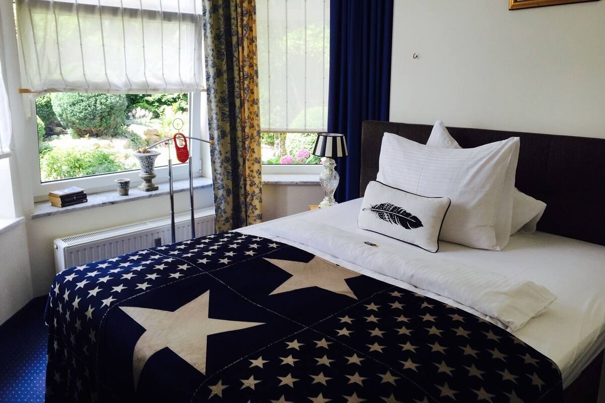 A neatly made bed with a star-patterned bedspread, a bedside lamp, and a window with curtains.