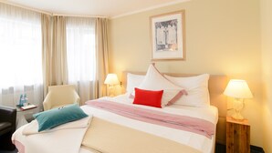 Habitación de hotel con cama, dos lámparas de noche, una silla, un escritorio con lámpara y una ventana con cortinas.