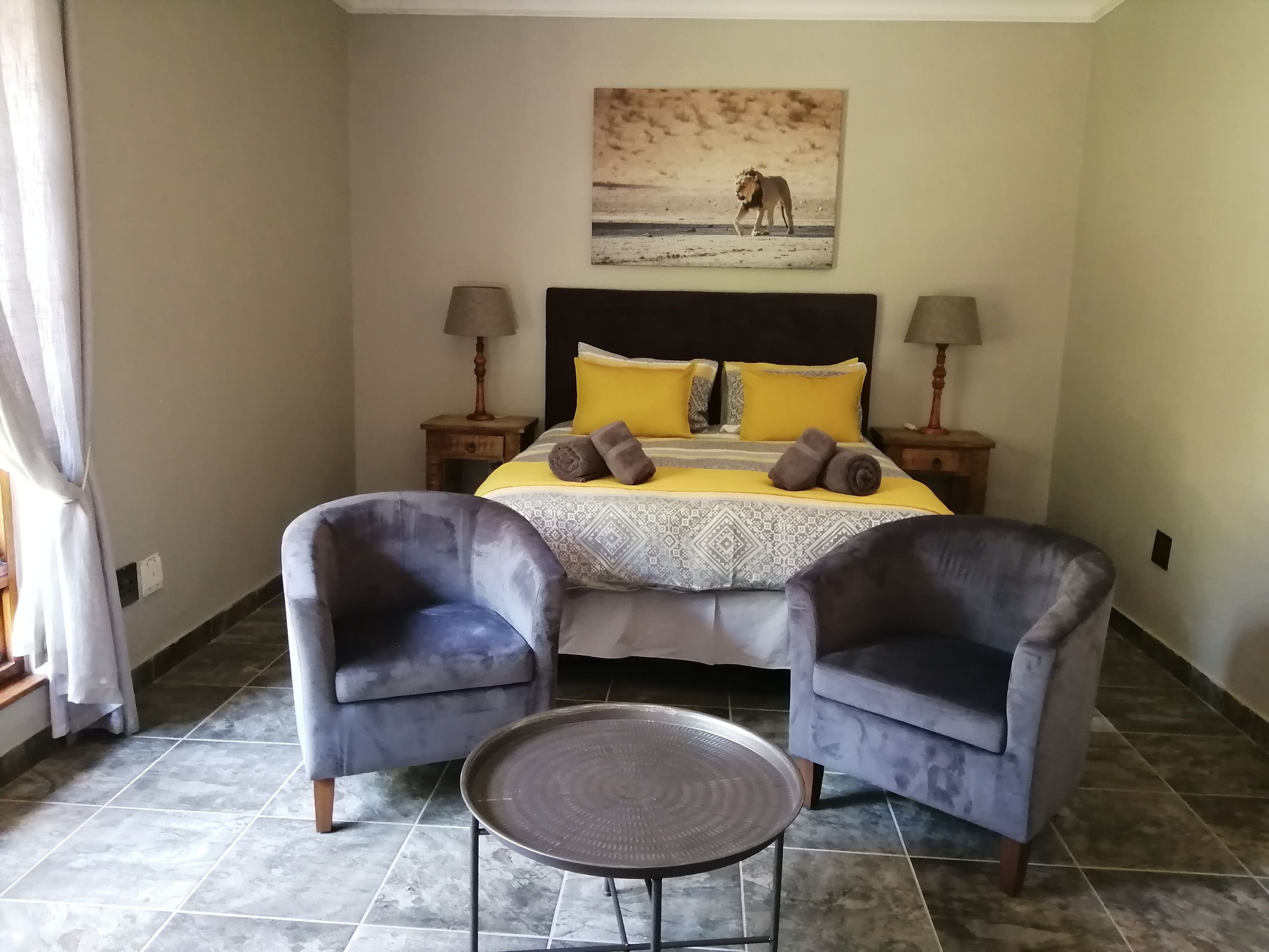 A bedroom with a bed, two armchairs, a small round table, and a painting of a lion on the wall.