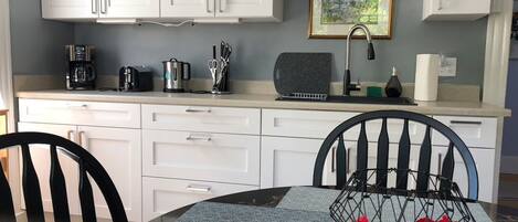 A modern kitchen with white cabinets, a coffee maker, a kettle, and a toaster. There is a framed picture on the wall and a vase with red flowers on the table.