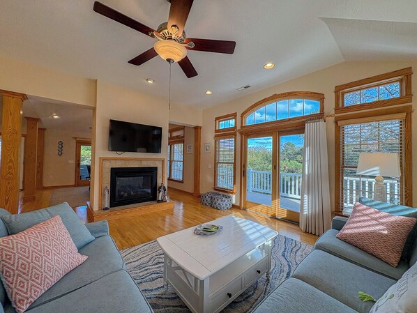 A spacious living room with a fireplace, a ceiling fan, and large windows offering a view of the outdoors.
