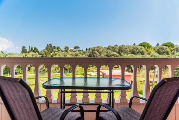 Un balcon avec une table en verre et deux chaises, donnant sur un jardin luxuriant et une piscine.