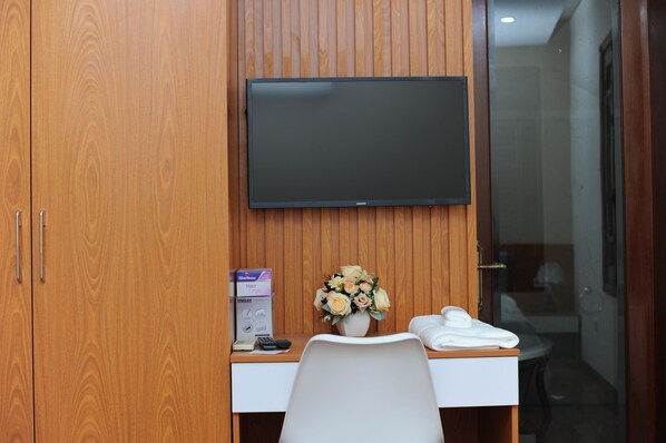 A hotel room with a wooden wardrobe, a desk with a vase of flowers, a flat-screen TV, and a white chair. - Quang Anh Hotel (Hanoi)