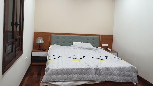 A bedroom with a bed, a nightstand, a lamp, an air conditioner, and a wooden door. - Quang Anh Hotel (Hanoi)