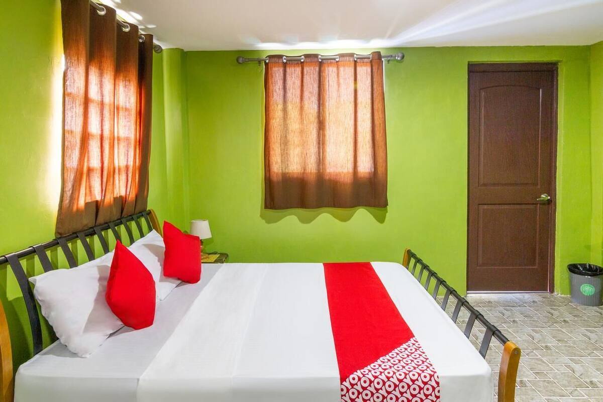 A hotel room with a bed, a nightstand, a lamp, a window with curtains, a door, and a trash bin.