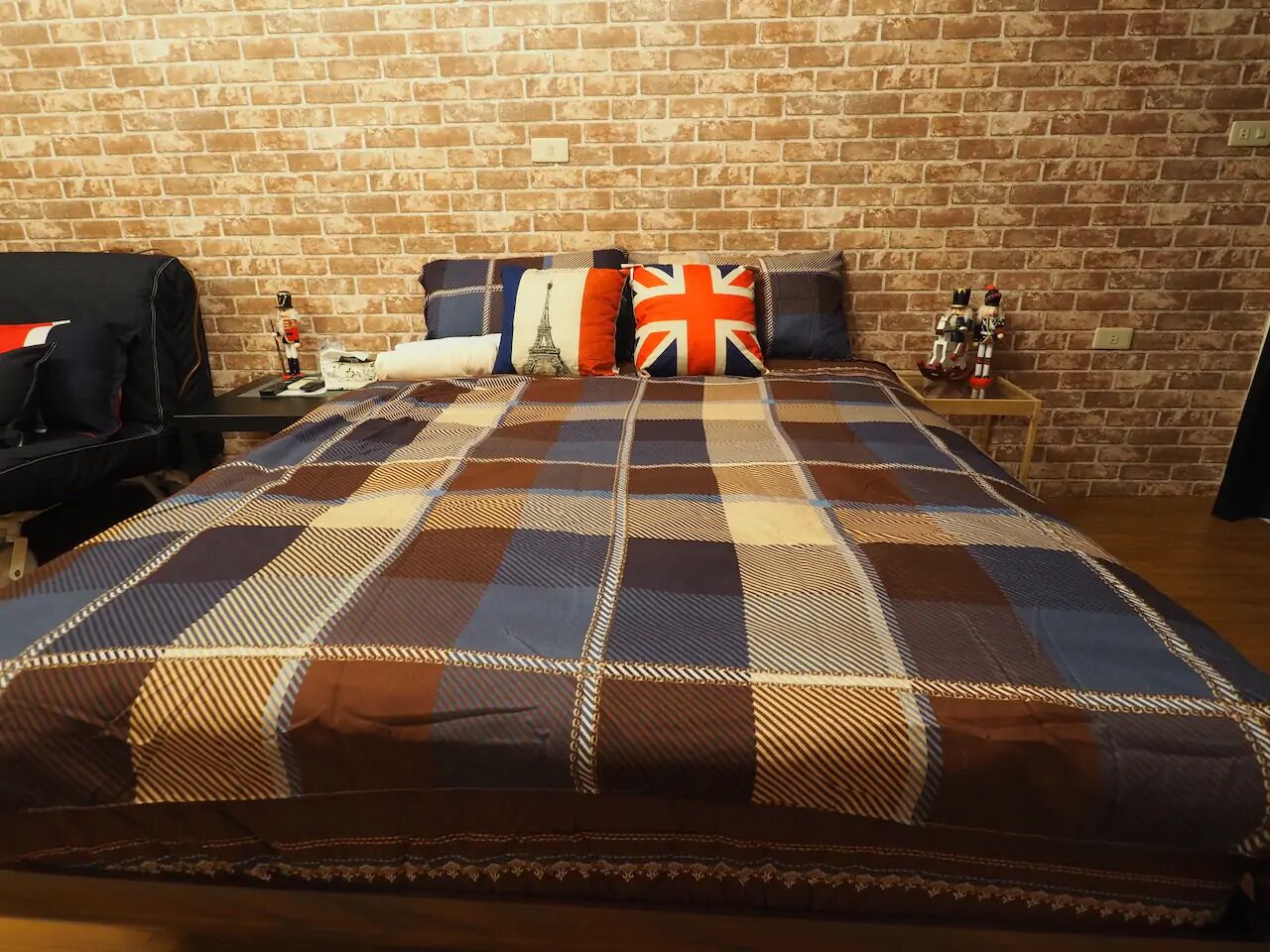 A bed with a plaid bedspread, a suitcase, and a small table with a bottle and a figurine.