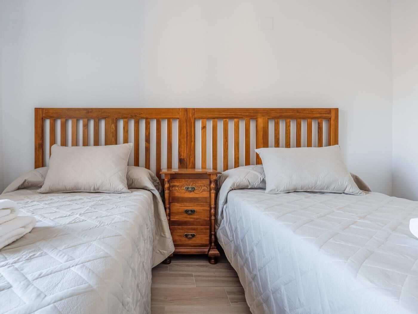 Una cama doble con cabecera de madera, una mesita de noche con cajones y una pared blanca lisa.