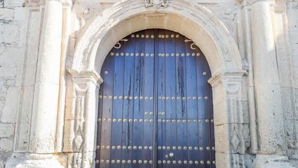 Une grande porte à deux battants, richement décorée, de couleur bleu foncé et ornée de clous métalliques, intégrée dans une structure architecturale classique aux sculptures détaillées.