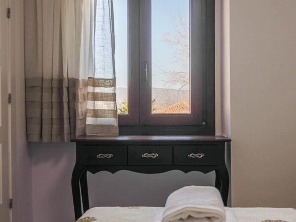 A bedroom with a bed, a window with curtains, a nightstand with drawers, and a view of a landscape outside.