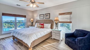 Ein Schlafzimmer mit einem großen Bett, einem Deckenventilator, einem Nachttisch, einem Sessel und einem Fenster mit Blick auf einen Pool und Palmen.