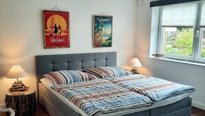 Un dormitorio con una cama tapizada en gris, ropa de cama a rayas, dos carteles colgados en la pared y una ventana con persianas.