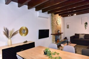 A modern living room with a wooden dining table, a black sofa, a stone fireplace, and a mounted TV.