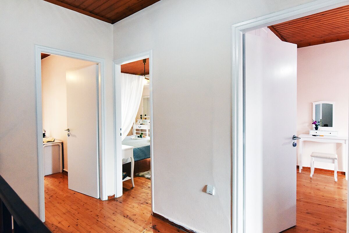 A hallway with wooden flooring, white walls, and a ceiling with exposed beams. There is a white desk with a chair and a mirror on the right side.