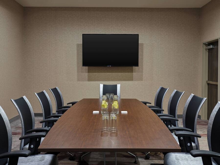 Un salón de conferencias con una larga mesa, sillas, un televisor de pantalla plana y un vaso de agua sobre la mesa.