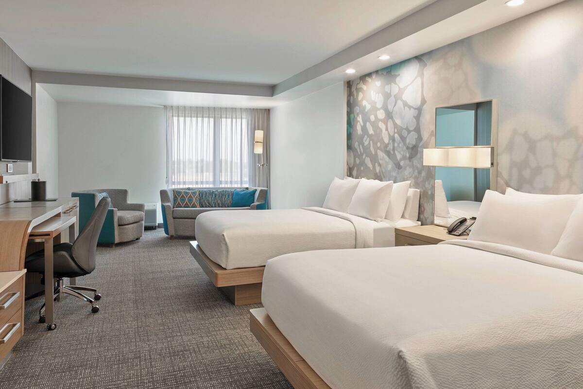 A hotel room with two beds, a desk, a chair, a lamp, and a TV.