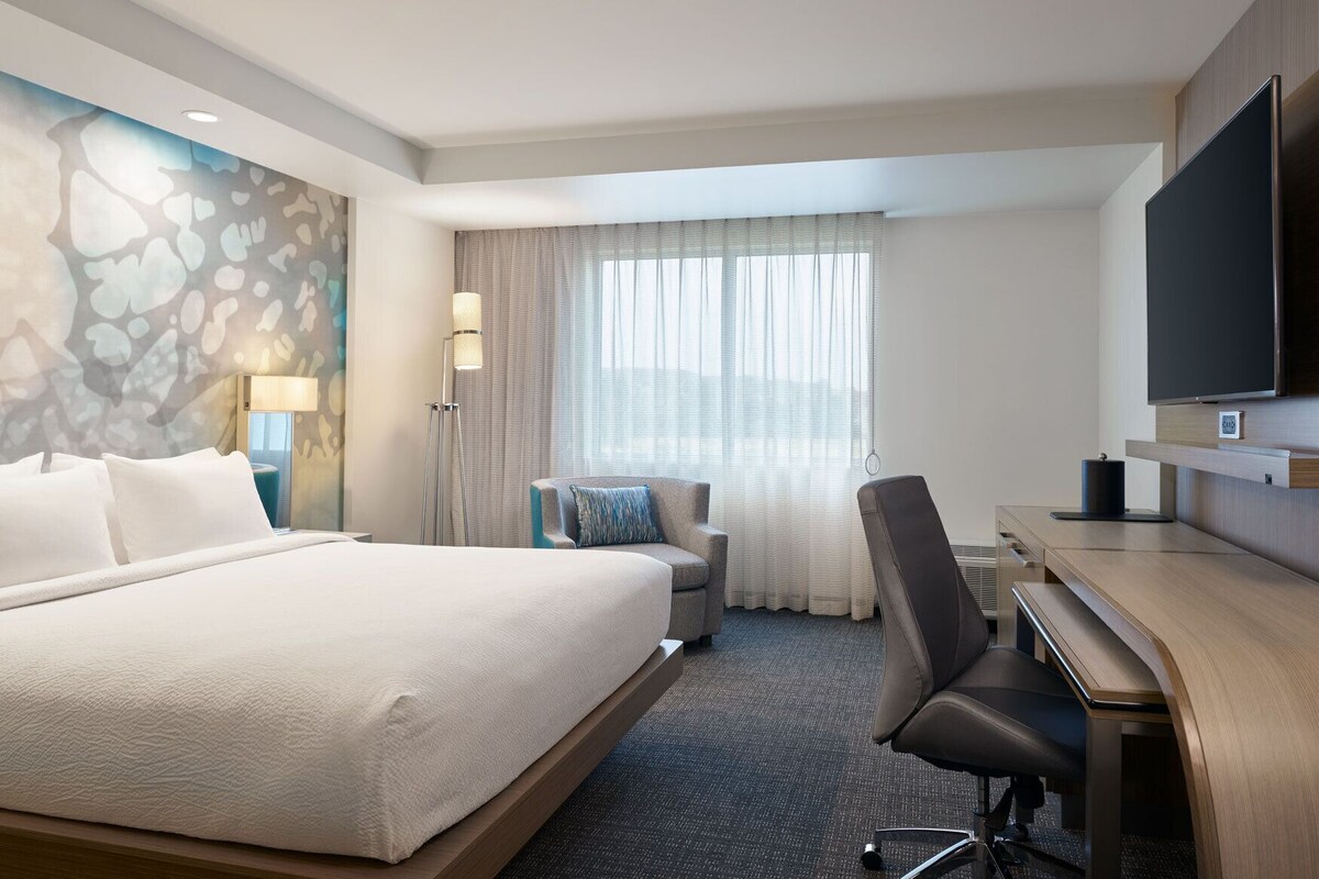 A hotel room with a large bed, a desk, a chair, a flat-screen TV, a lamp, and a window with curtains.
