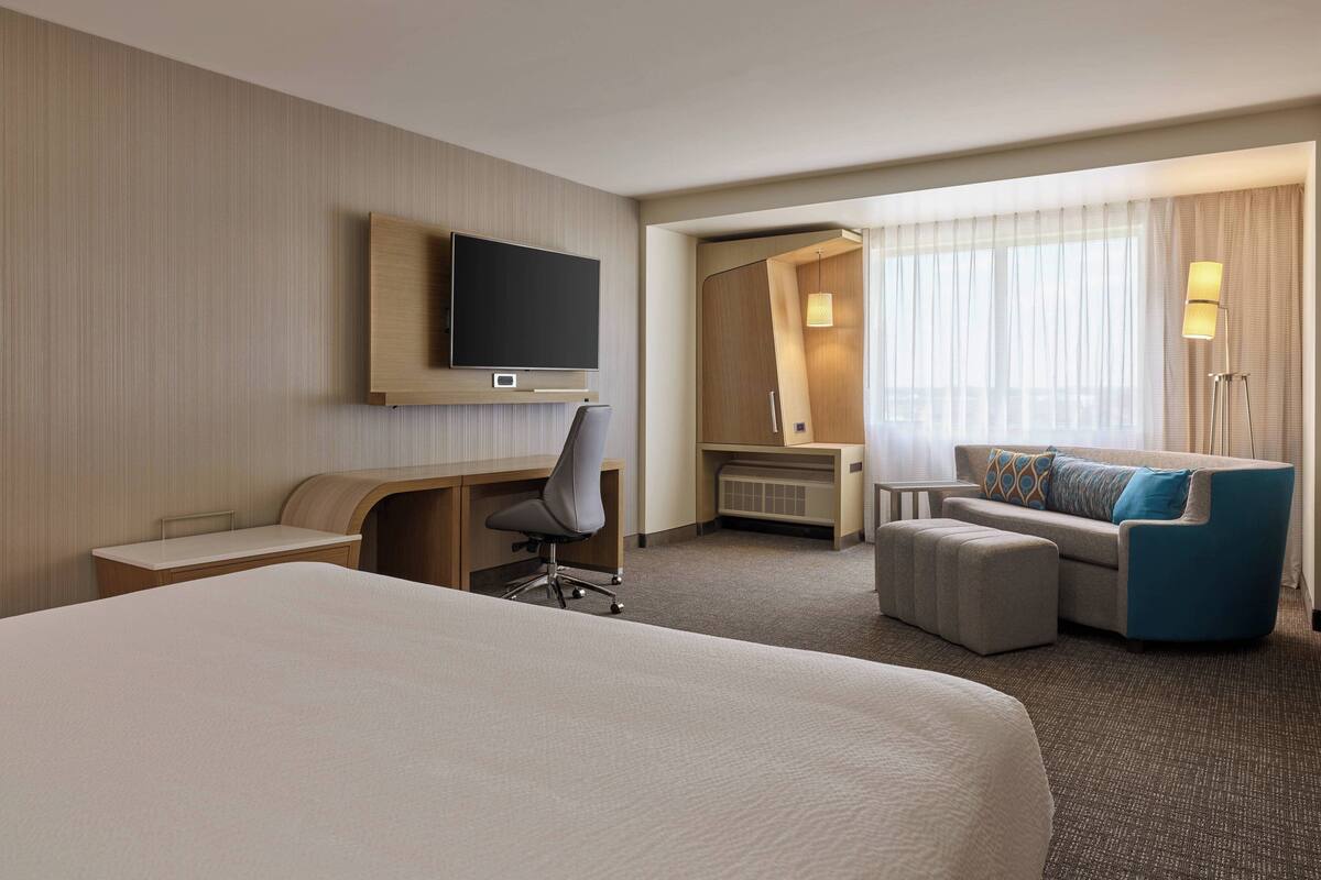 A hotel room with a large bed, a desk with a chair, a sofa, a television, and a window with curtains.