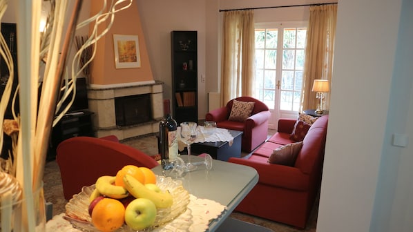 A cozy living room with a fireplace, red sofas, a dining table with fruit, and a bookshelf.